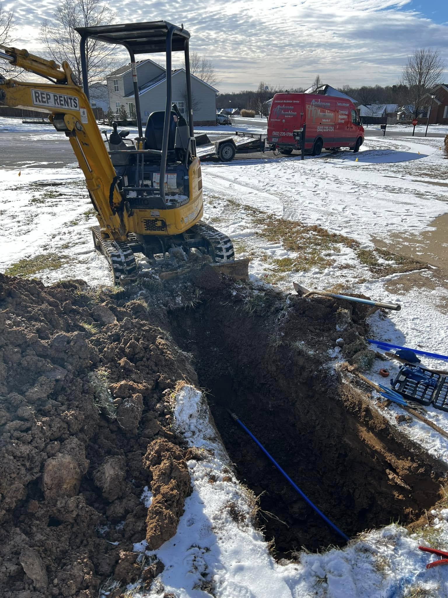 Front yard with open 3 foot by 8 foot trench. A backhoe owned by the equipment rental company REP Rents is positioned at the far end of the trench. A new blue water line is visible, which replaces a leaking section of an old underground water line, connected by brass fittings. Some tools are visible on the lawn to the right and at the near end of the trench. An Anderson Plumbing is visible on the street in the background. Three neighbor houses are visible in the background of a spacious, newer development, with a wooded area in the far background, 400 yards away.