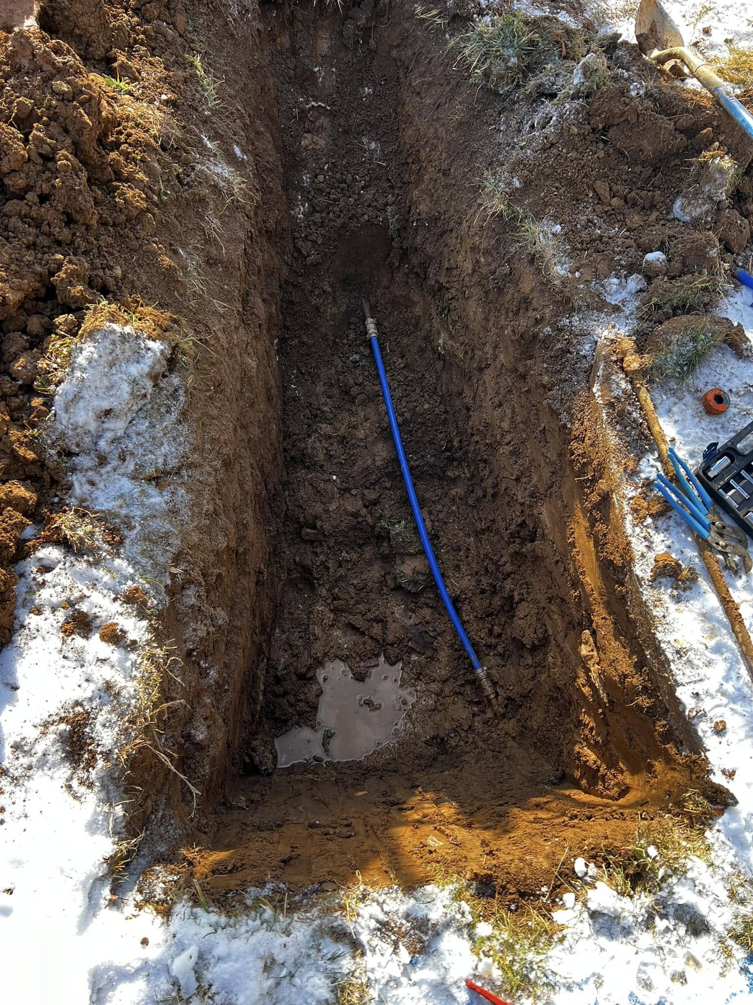 Open 3 foot by 8 foot trench with new blue water line that replaces old, leaking section with a brass connector at each end.