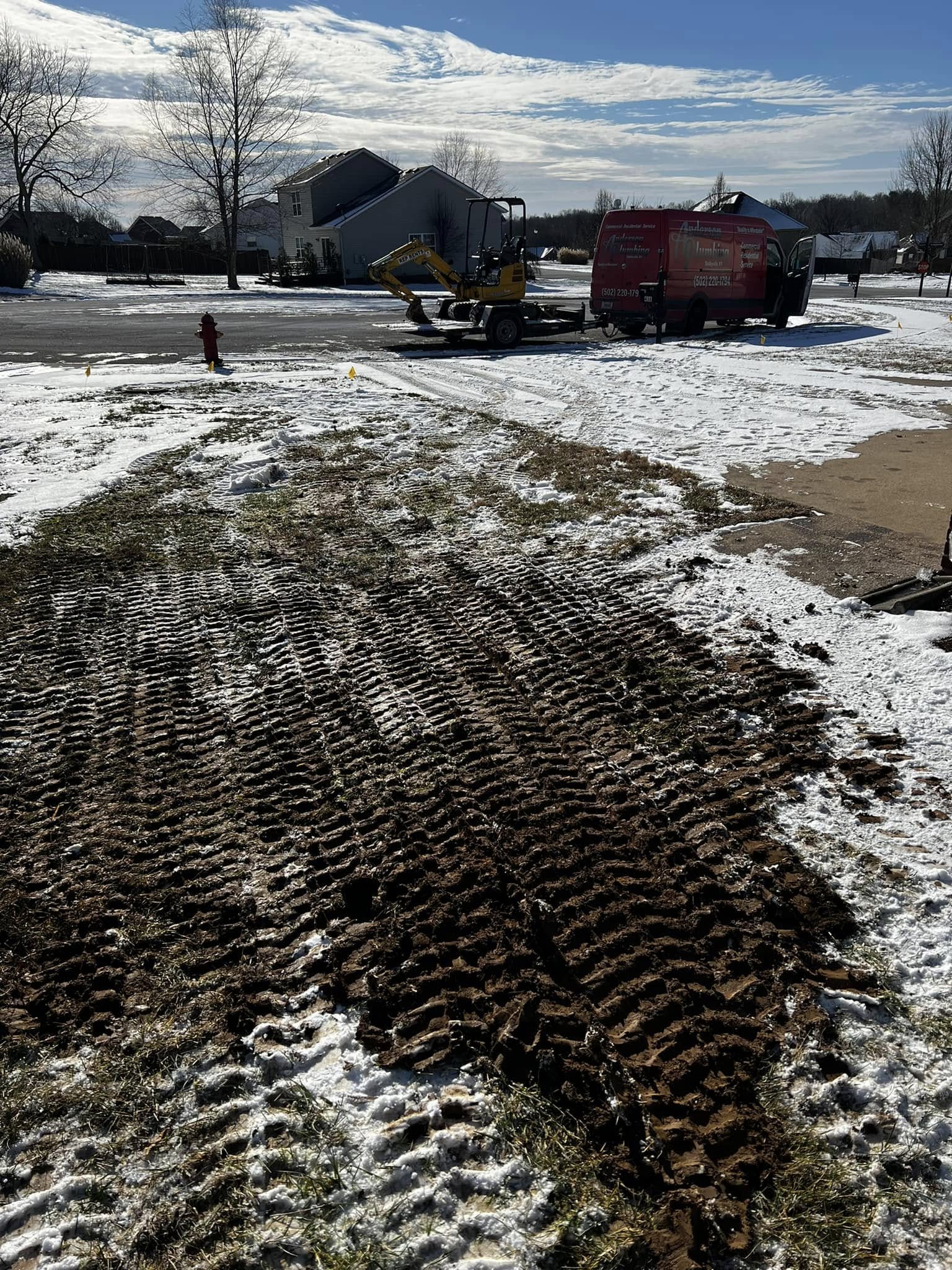 Front yard with backhoe in background, after completing underground water leak repair. Small caution flag markers visible where utility companies have marked off location of other underground service lines. There is a thin layer of snow on the ground.